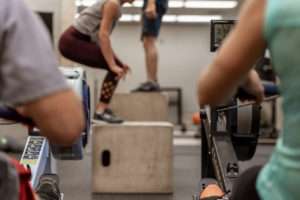 two people working out on rowing machine with others doing box jumps in front of them